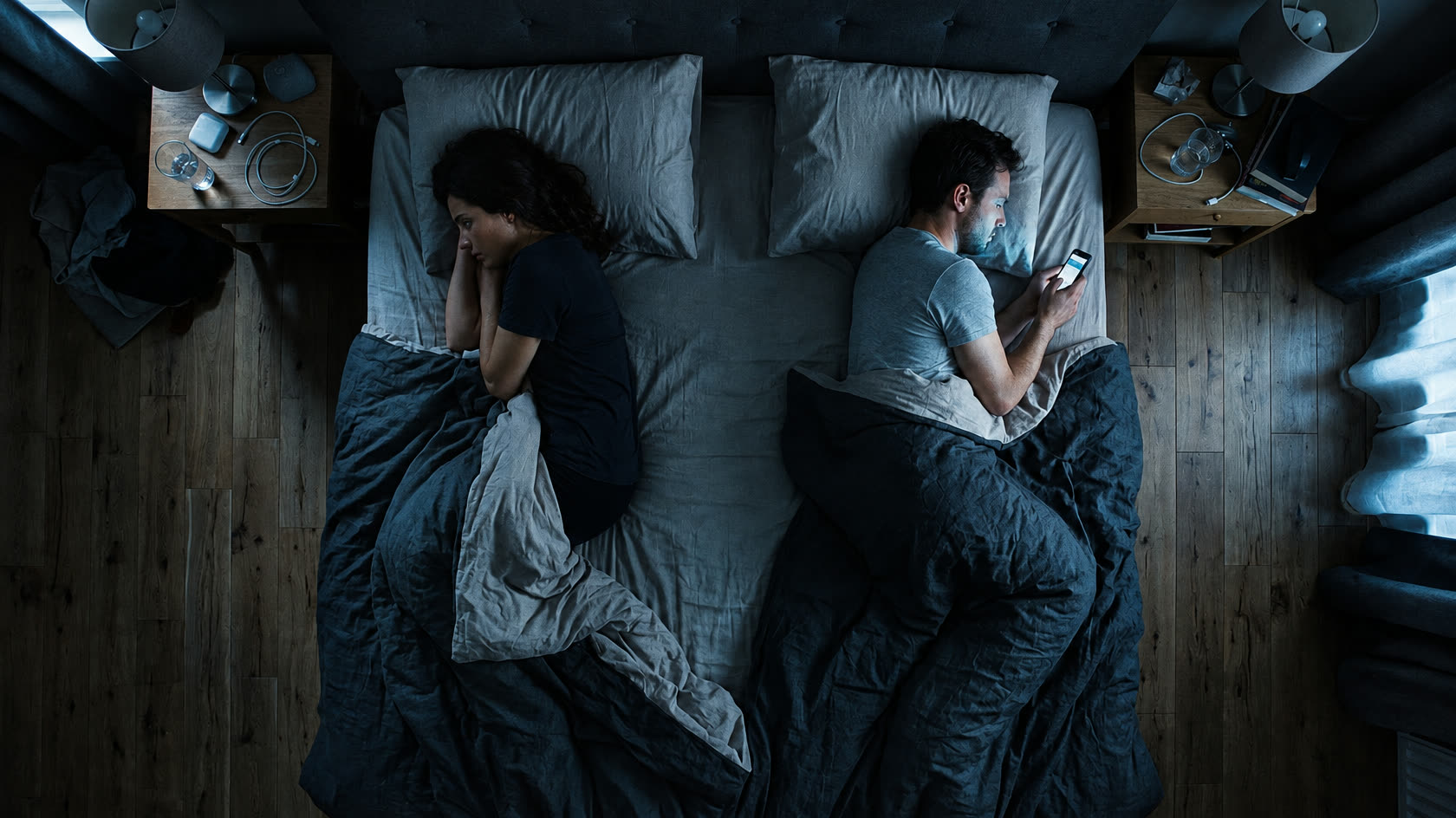 A couple in bed at night seen from above, backs turned. One is lit by a phone.